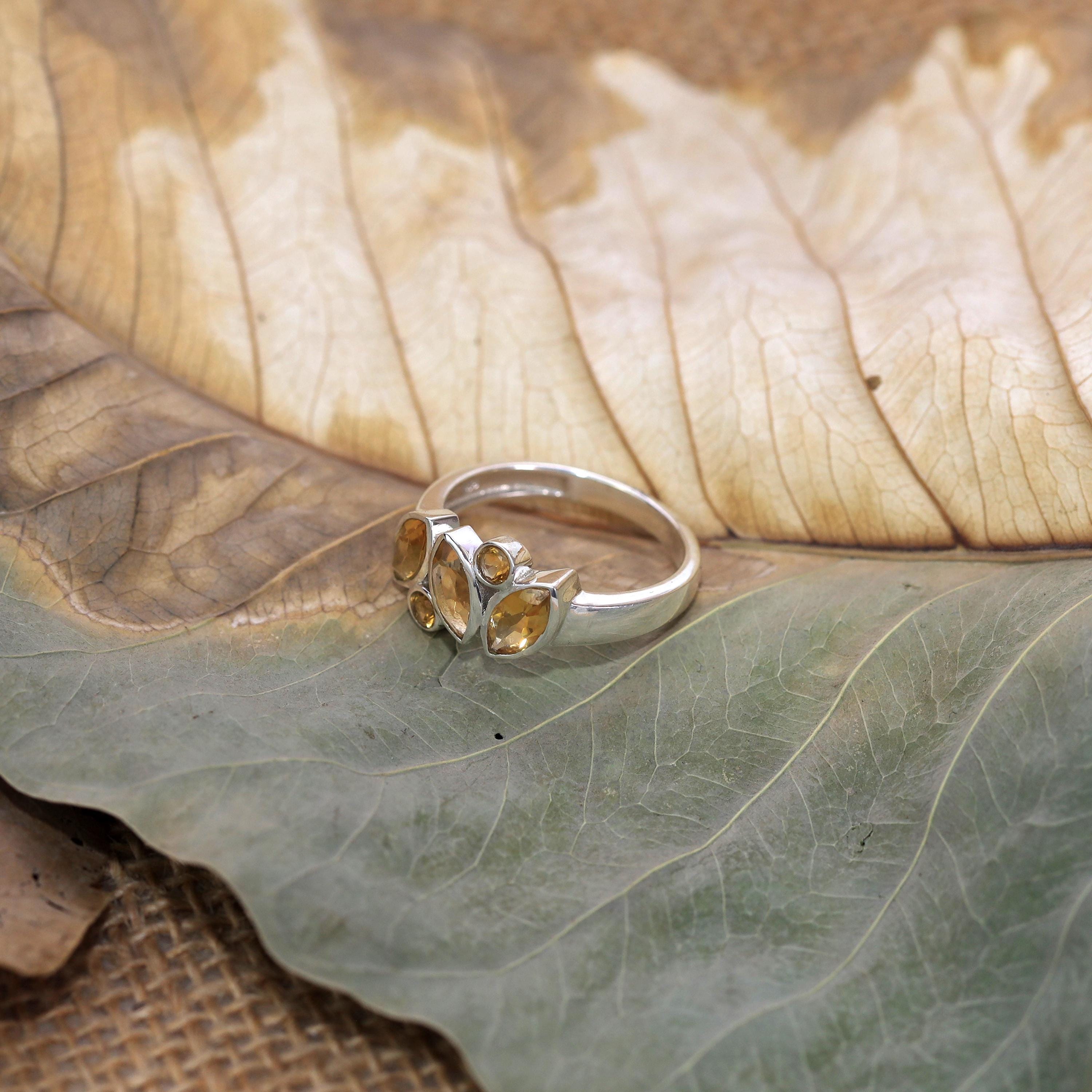 May include: A silver ring adorned with a cluster of amber-coloured, oval gemstones. The ring is displayed on a dried leaf, highlighting the jewellery's design and colour.