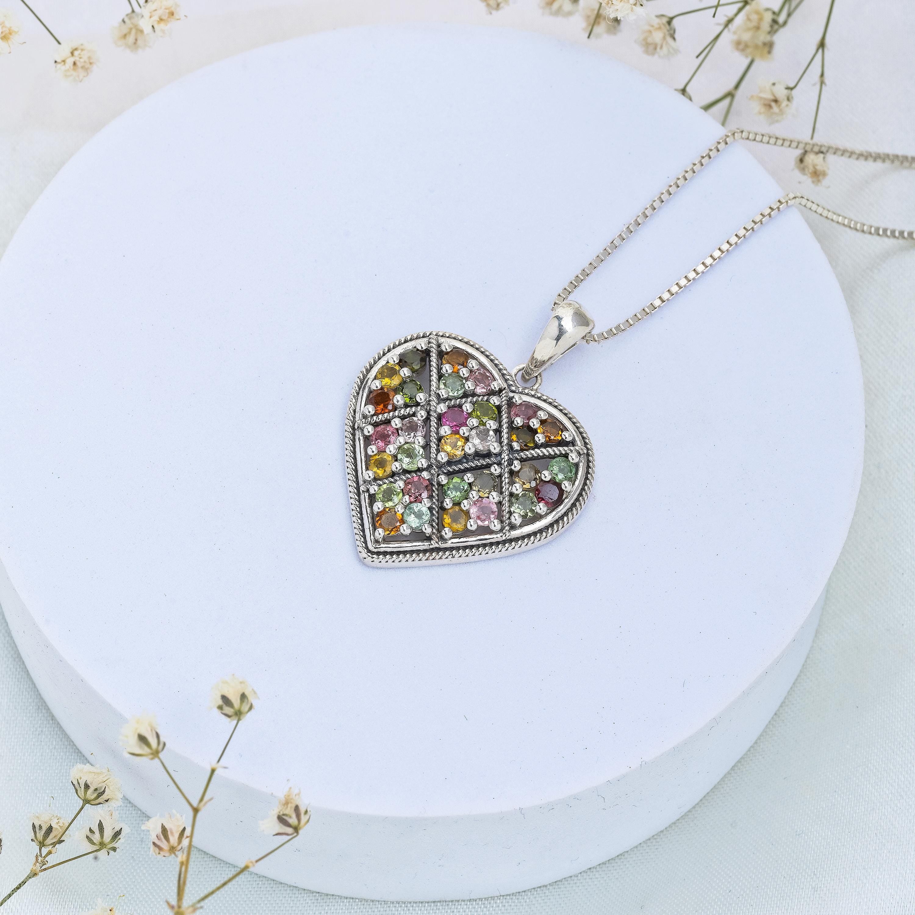 May include: A silver heart-shaped pendant necklace with a detailed design and a variety of colourful gemstones. The necklace is on a white background.